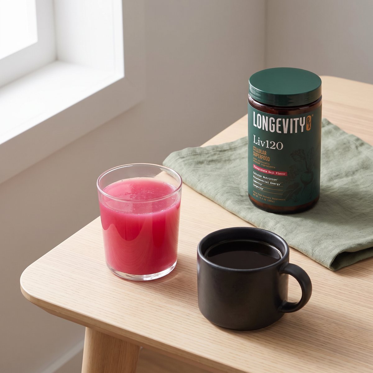 A glass of pink Liv120 next to a mug of black coffee, with the Liv120 jar behind