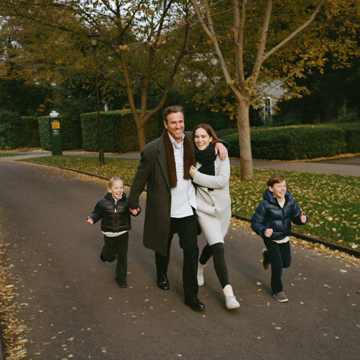 A smiling man in his sixties walking arm in arm with his adult daughter down a leafy street in late autumn, grandkids running ahead