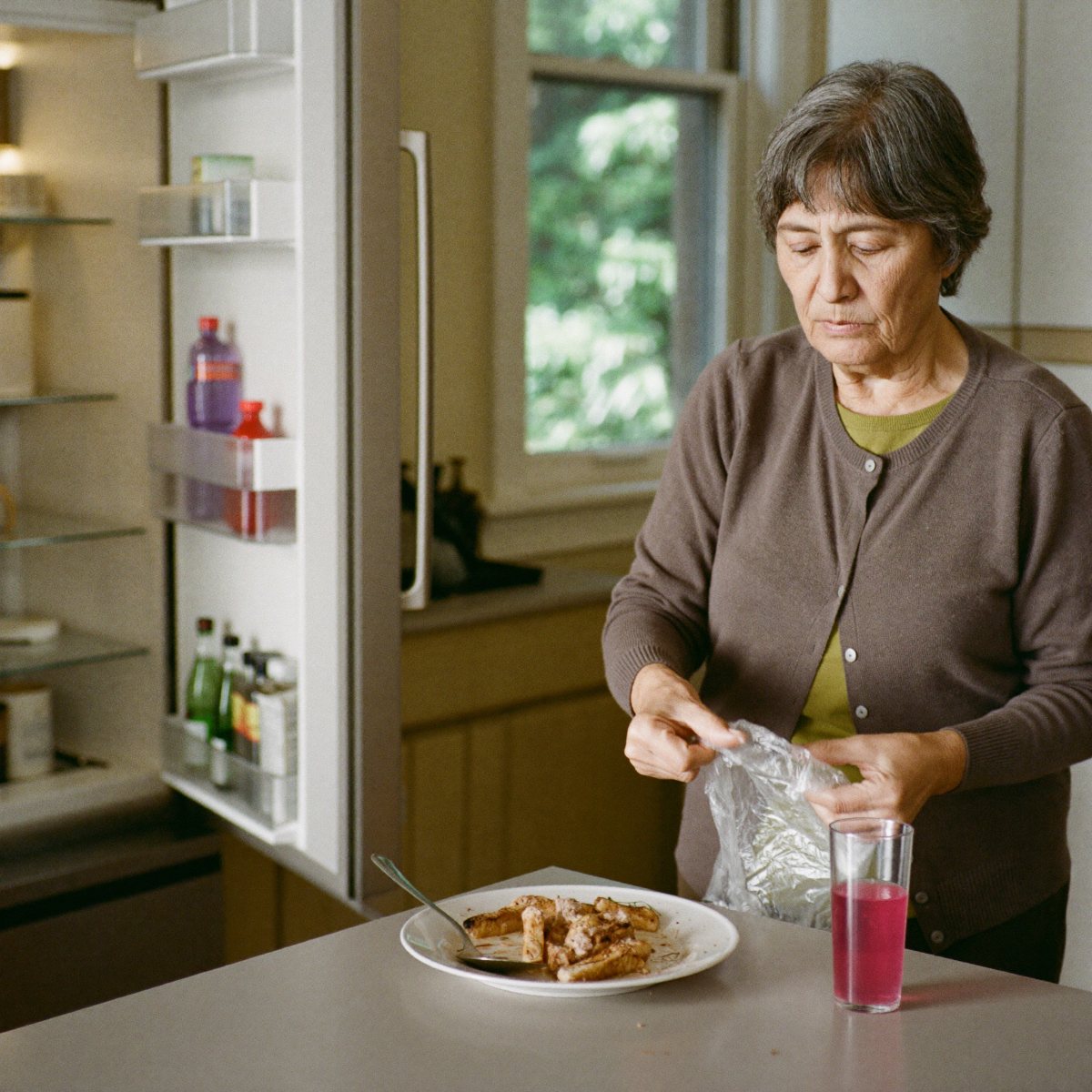 A woman in her sixties covering a half-eaten dinner plate with wrap before putting it in the fridge, a glass of pink drink on the counter